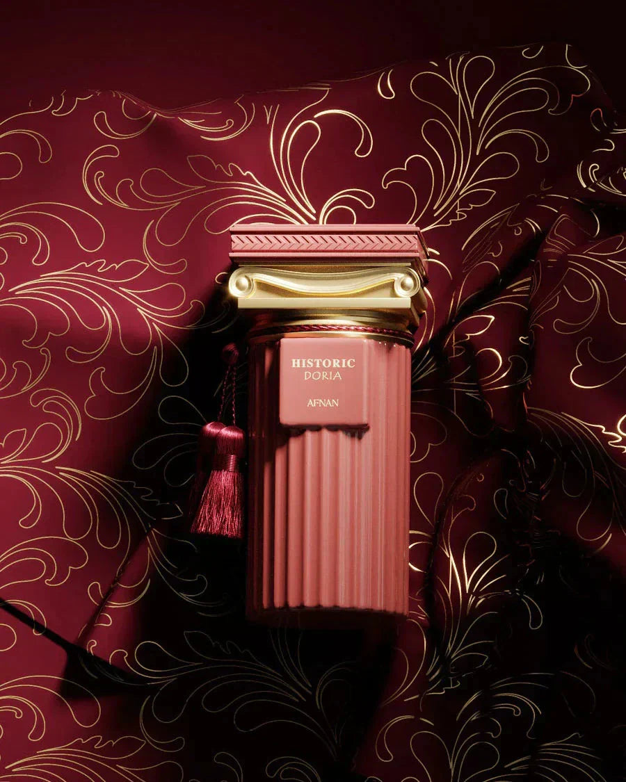 The image features an opulent perfume bottle against a richly patterned background. The bottle has a sophisticated design with a deep rosy pink hue and ribbed texture, crowned with a luxurious golden cap that has classical detailing. A matching rosy pink tassel hangs from the cap, adding to the elegance. The label on the bottle reads "HISTORIC DORIA AFNAN". The backdrop is a dark red fabric adorned with golden, swirling floral patterns that give the impression of luxury and exclusivity. 