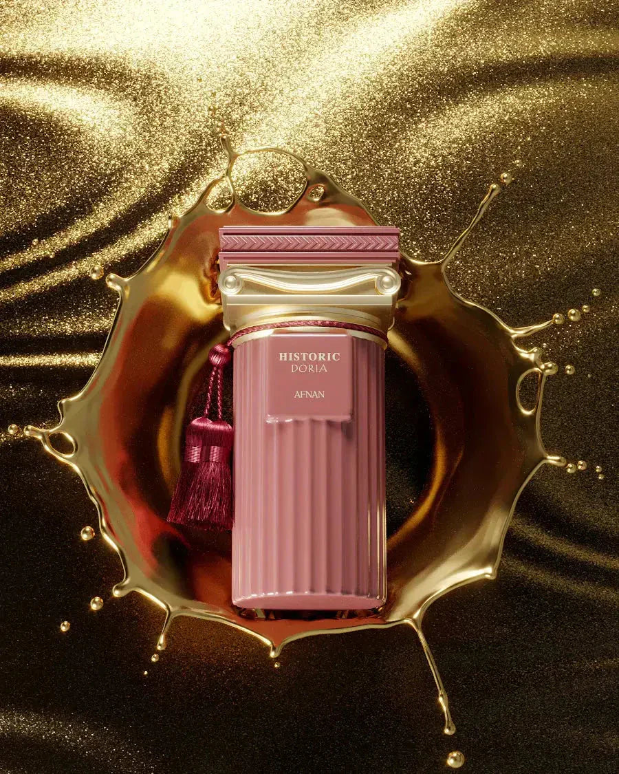 The image showcases a luxurious perfume bottle from the Afnan "Historic Doria" collection against a shimmering gold background with a dynamic splash of golden liquid surrounding it. The perfume bottle is a sophisticated pink with a ribbed texture, reminiscent of classical columns. A deep pink tassel adds a touch of opulence to the presentation. The bottle is labeled with "HISTORIC DORIA" and the Afnan brand name in an elegant font.