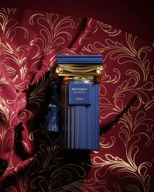 The image features a luxurious fragrance bottle placed against a richly patterned background. The bottle has a deep blue color with vertical ridges and is adorned with a gold cap that has a classical design, complete with a blue tassel. The gold label on the bottle reads "HISTORIC OLMEDA AFNAN." The backdrop is a dark red fabric with a glossy finish, embellished with a gold floral pattern that exudes opulence and elegance.