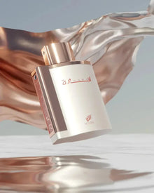 The image features an elegant perfume bottle in a dynamic setting. The bottle is matte white with a rose gold cap and details. The name "?????" is written on the front in red Arabic script, and the "AFNAN" logo is also in red below it. The bottle appears as if it is floating, with a fluid, shimmering rose gold fabric-like structure undulating in the background, creating a luxurious and dreamlike atmosphere.