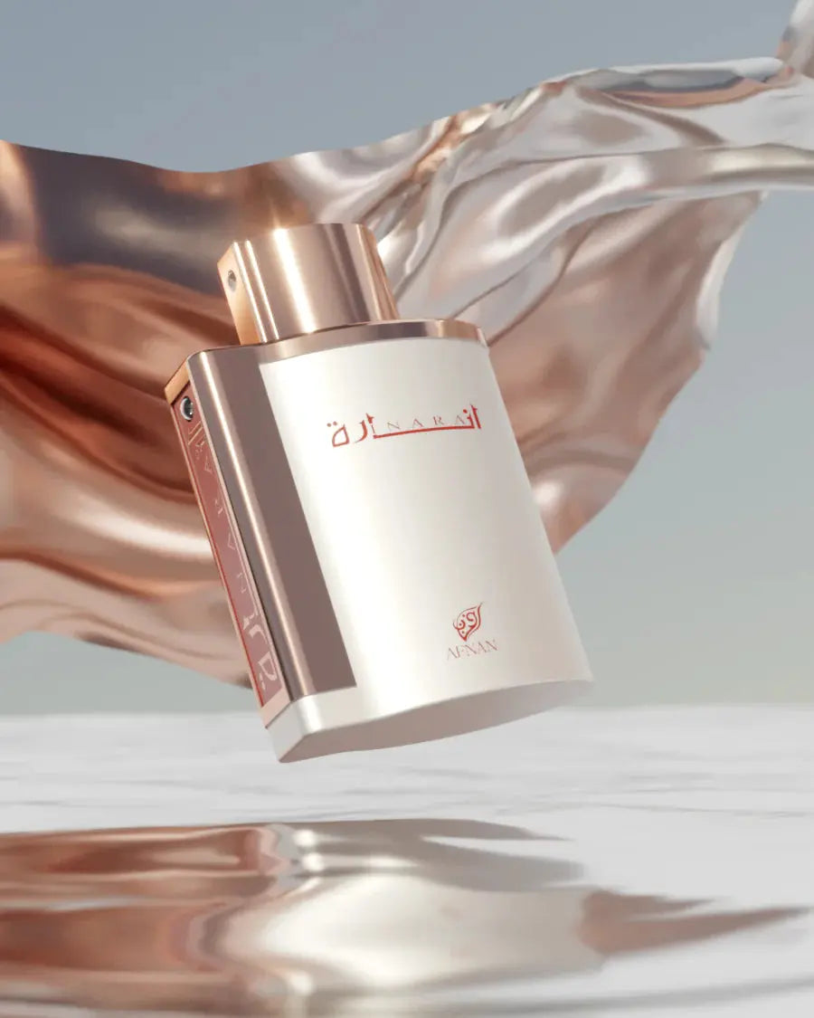 The image features an elegant perfume bottle in a dynamic setting. The bottle is matte white with a rose gold cap and details. The name "إنارة" is written on the front in red Arabic script, and the "AFNAN" logo is also in red below it. The bottle appears as if it is floating, with a fluid, shimmering rose gold fabric-like structure undulating in the background, creating a luxurious and dreamlike atmosphere.