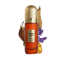 This image features a tall, cylindrical perfume bottle with a gold cap, labeled "Jamaican Tobacco" by Ibraheem Al Qurashi. The bottle is amber-colored with a textured surface, and it shows the volume as "100 ml / 3.4 fl.oz." A dry tobacco leaf, some lavender sprigs, and pieces of amber resin are placed around the bottle, suggesting the scent notes of the fragrance.