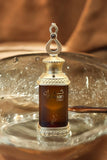 This image features a perfume bottle labeled "Kayaan Gold" resting on a glass surface. The bottle has a regal design with a gold-toned cap adorned with decorative elements and a loop on top, suggesting it can be used as a pendant or for easy carrying. The dark amber liquid inside indicates the perfume might have a rich and possibly woody or oriental fragrance.
