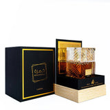 This image showcases a perfume set by Lattafa, featuring the KHAMRAH fragrance. It includes a square, crystal-cut glass perfume bottle, with golden-yellow liquid visible inside, placed on a pedestal-like platform with a golden front panel. The pedestal al