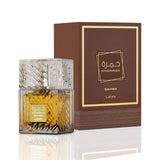 This image features a square crystal-cut perfume bottle with golden-yellow liquid, labeled "KHAMRAH QAHWA" by Lattafa, in front of its packaging. The perfume bottle showcases intricate glasswork that reflects the light, and a clear, matching glass cap. The front of the bottle has a golden label with the perfume name in both Arabic and English, alongside the Lattafa brand name. The packaging behind the bottle is a book-style box with a rich brown color, a textured appearance resembling a leather spine.