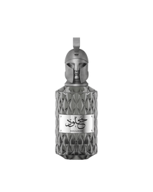 A product shot of Le Falcone Niche Collection’s “Khulood” perfume bottle on a white background: a tall, amber-colored glass vessel with a diamond-cut texture and a polished bronze plaque engraved with Arabic calligraphy above the word “Khulood.” The bottle is crowned by an ornate bronze cap sculpted into a stylized warrior’s helmet with a crest-style loop at the top, conveying a luxurious, sculptural aesthetic.