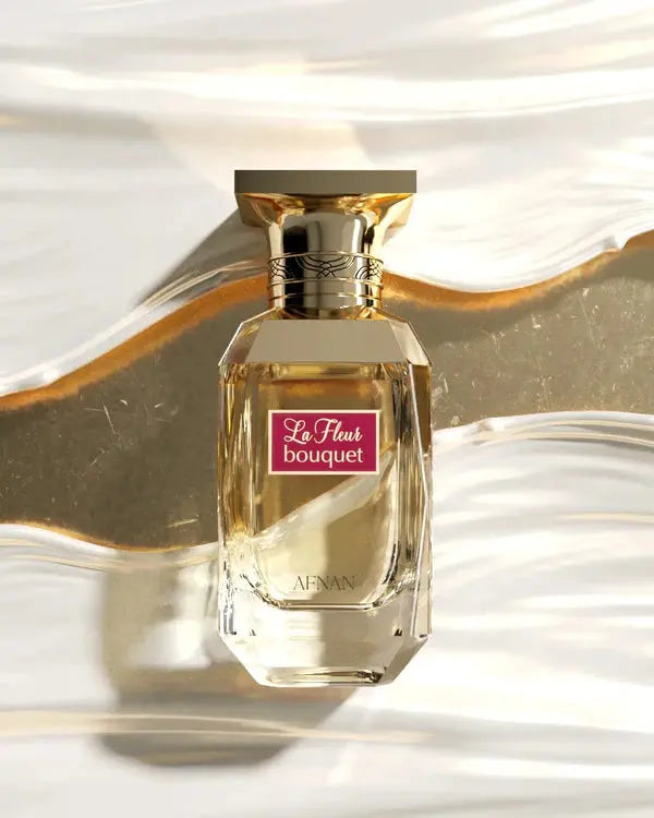 The image depicts a luxurious perfume bottle set against a backdrop that mimics flowing golden fabric. The bottle is made of clear glass with a geometric design that elegantly refracts light, enhancing its premium look. It has a square golden cap with intricate line engravings around the collar. The label on the bottle is maroon with the words "La Fleur bouquet" written in sophisticated white cursive script, and the brand name "AFNAN" is positioned on the lower part of the bottle in gold to match the cap.