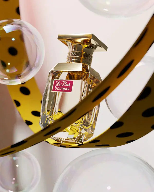 The image displays an artistic perfume bottle presentation. The bottle is transparent with a multifaceted glass design that catches the light, filled with golden-colored perfume. It features a square golden cap with a decorative pattern around the neck. The front of the bottle has a maroon label with "La Fleur bouquet" in white cursive font, and the "AFNAN" logo is present at the bottom. The bottle is framed by abstract golden bands with cut-out polka dots.