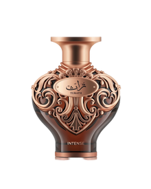 A high-resolution product shot of Le Falcone Niche Collection’s “Turath Intense” perfume bottle against a white background. The bottle has a dark amber, heart-shaped glass body topped with a wide, copper-toned neck and cap. Its upper half is covered in ornate, scrolling filigree in a polished copper finish, forming elegant arabesque patterns. A matching copper plaque at the front bears Arabic calligraphy above the word “TURATH,” and “INTENSE” appears in gold lettering near the base.