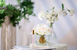 The image showcases an elegant perfume presentation on a marble table, framed by a soft-focus floral background. In the foreground, a clear glass vase holds a large, beautifully detailed pale pink rose, adorned with a gold label reading "Cote Noire." Beside the vase, there is a slender perfume roller with a clear body and gold cap, labeled "Petale De Rose." In the background, a small glass vase containing white blossoms adds a delicate contrast.