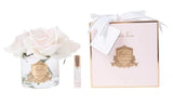 The image features a charming and sophisticated Cote Noire product display on a white background. On the left, there's a clear glass vase containing several pale pink roses, with a gold label enhancing its elegance. Next to the vase is a small perfume roller with a clear body and a gold cap. On the right, a light pink gift box with a gold crest and a white ribbon tied in a bow holds the focus, accompanied by a gift tag.