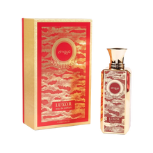 The image shows a bottle of "Luxor" eau de parfum by Zimaya alongside its packaging. The perfume bottle is tall and rectangular with a gold cap, featuring an ornate red and gold label with the brand name "zimaya" in a circular emblem near the top. The word "Luxor" is written in bold at the bottom of the label, along with "eau de parfum." The packaging box matches the bottle's design, with a red and gold pattern and similar branding elements.