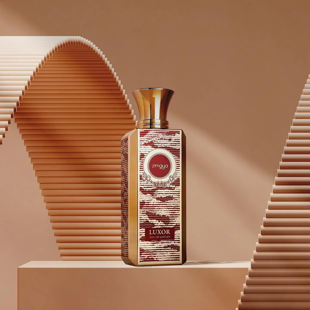 The image shows a bottle of "Luxor" eau de parfum by Zimaya. The perfume bottle is tall and rectangular with a gold cap. The label features intricate designs in red and gold, with the brand name "zimaya" in a circular emblem near the top. The word "Luxor" is written in bold at the bottom of the label, along with "eau de parfum." The background consists of a sophisticated, abstract design with beige tones and curved, ribbed structures, enhancing the luxurious and elegant presentation of the product.