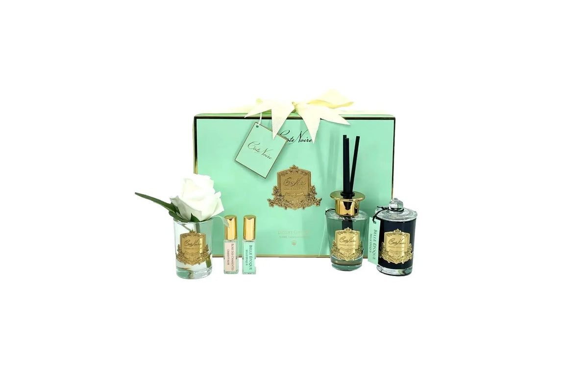 A luxury gift set featuring various scented products from Cote Noire. The set includes a glass jar with a white rose, three small perfume vials, a reed diffuser, and a scented candle, all adorned with gold labels. The items are elegantly arranged in front of a mint green box with gold accents and a cream-colored ribbon.