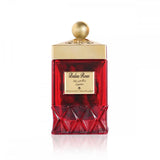 The image features a luxurious perfume bottle. The bottle has a rich, deep red color with a faceted geometric design at the bottom, giving it a crystal-like appearance. The top half of the bottle is smooth, and it has a gold-colored label with the words "Balas Rose" and Arabic script below it, suggesting the fragrance name and possibly the brand. Above the label, the neck and cap of the bottle are gold, with the cap adorned by intricate detailing and topped with a spherical golden knob. 
