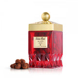 The image shows an elegant perfume bottle. The bottle has a square base with a faceted, diamond-cut design in a vivid red color, giving it a rich, luxurious look. The upper section of the bottle is smooth with a gold label that has "Balas Rose" in English and Arabic script. The cap is golden with a textured design, topped with a spherical knob. Next to the bottle, there is a small cluster of dark brown, textured balls, possibly scented bakhoor.
