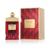 The image features a luxurious fragrance product with its packaging on a white background. On the left side is a perfume bottle with a deep red, faceted base resembling cut gemstones, and a smooth upper portion with a gold label front and center. The label displays "Balas Rose" in English and Arabic script, suggesting the fragrance name. The bottle is topped with a golden cap adorned with a texture resembling small beads, crowned by a spherical knob.