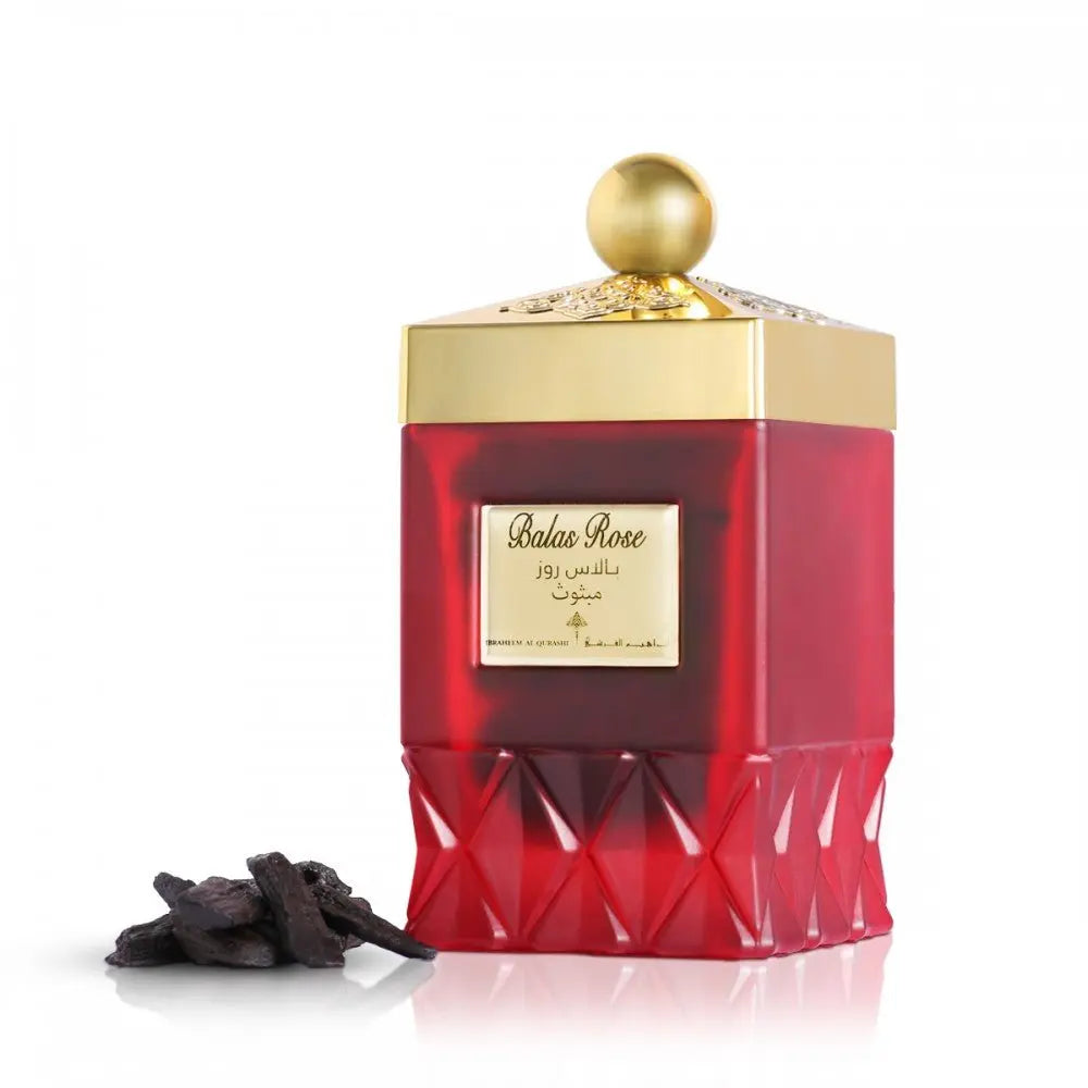 The photo features an elegant perfume bottle with a rich, translucent red color and a faceted design at the base. The bottle is adorned with a gold cap that has intricate patterns and a simple spherical knob on top. A label on the bottle displays "Balas Rose" in English and Arabic script, along with the brand name "Ibraheem Al Qurashi". Beside the bottle, there is a small cluster of dark, resinous pieces, possibly oud or another aromatic substance.