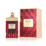 The image displays a striking red perfume bottle with a textured base alongside its packaging. The bottle features a golden cap with an ornate design and a label that reads "Balas Rose" in both English and Arabic script, with the brand "Ibraheem Al Qurashi" beneath it. The accompanying box shares the bottle's red and gold color scheme with a diamond pattern that complements the bottle's design. 