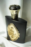 The image shows an ornately designed perfume bottle with a crown-shaped cap, placed on top of a matte black box. The cap is a reflective gold color, intricately detailed with a textured pattern and traditional embellishments, which give it a royal and luxurious appearance. On the front of the black box is a gold label with elaborate scrollwork and Arabic calligraphy, which indicates the fragrance's name or brand, suggesting that the perfume is of Middle Eastern origin. 