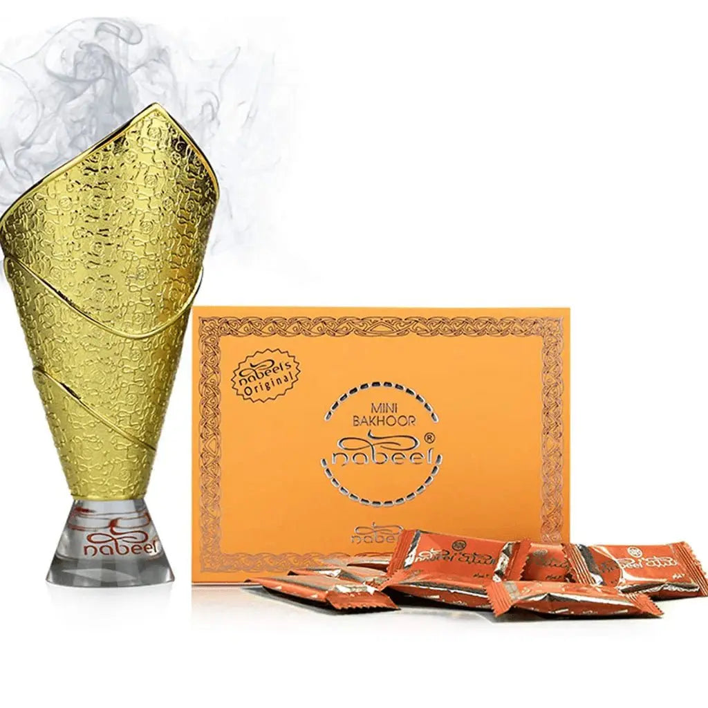The image displays a textured golden bakhoor burner emitting smoke, indicating its use, set on a metallic silver base with the "nabeel" brand name. In the background, there is a flat, orange box labeled "MINI BAKHOOR nabeel®" with intricate lace-like patterns around the edges and the brand logo in the center. Scattered in front of the box are several small packets with a matching orange color and the "Nabeel" brand name, which contain the bakhoor.