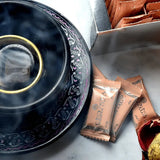 The image shows a close-up view of a black bakhoor burner with decorative patterns around its perimeter, and a golden bowl inside from which smoke is rising, suggesting that bakhoor is being burned. In the foreground, there are a few small, sealed packets in shades of orange and brown with the "Nabeel" brand name and logo, presumably containing bakhoor. 