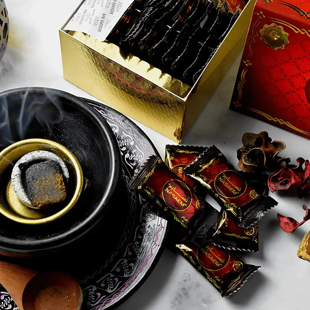 The image shows an arrangement of bakhoor-related items. In the foreground, there's a traditional black bakhoor burner with a golden bowl inside, containing a lit piece of bakhoor emitting smoke. Around the burner are several small, sealed packets with a maroon and gold color scheme and the "Nasaem" brand name, which likely contain bakhoor. In the background, a gold-colored box with a reflective surface is open, displaying a row of the same bakhoor packets.