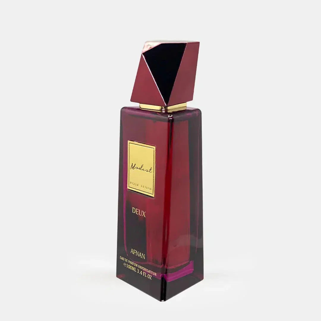 he image showcases "Modest Deux Pour Femme" by Afnan Perfumes, a perfume bottle with a rich, burgundy-hued glass body and an angular, geometric cap with a reflective golden finish. A gold label on the front features the perfume's name in an elegant font, and the bottom of the bottle includes the essential details of the fragrance's size and concentration. 