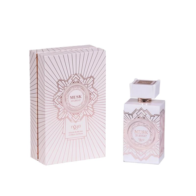 The image shows a bottle of "Musk Is Great" extrait de parfum by Noya alongside its packaging. The perfume bottle is rectangular and white with intricate geometric designs in a metallic rose gold color. It has a distinctive, square-shaped cap with vertical ridges and a rose gold accent at the base. The front of the bottle features a circular label with the text "Musk Is Great" and the brand name "Noya." The matching packaging box is also white with metallic rose gold geometric patterns.
