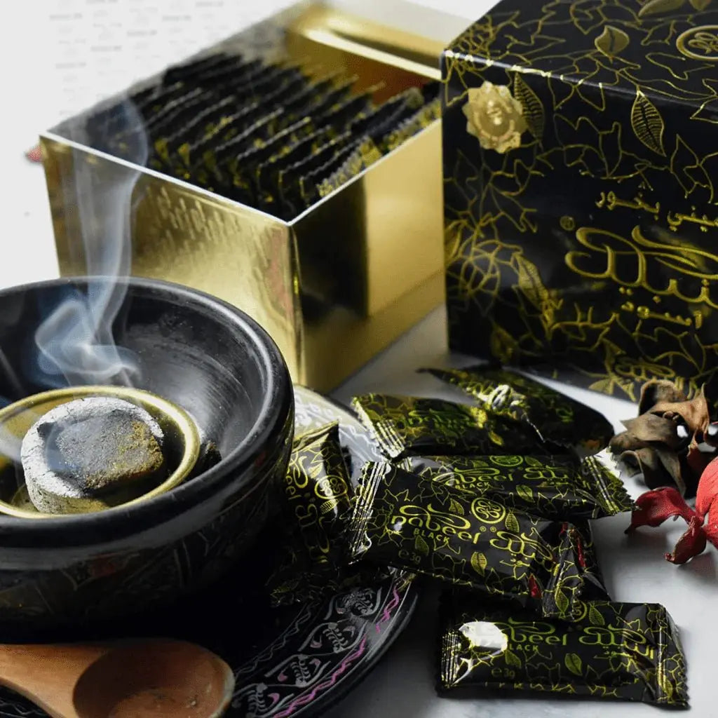 The image depicts a traditional setting where bakhoor, an aromatic product, is being used. In the foreground is a black bakhoor burner with a lit piece inside, from which smoke is rising, indicating that the bakhoor is being burned for fragrance. Surrounding the burner are several individually wrapped packets with a black and gold pattern, labeled "Nabeel", which likely contain the bakhoor. To the right, a gold-colored box is partially open, revealing rows of the same bakhoor packets.