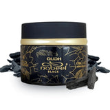 The image shows a black cylindrical container of "OUDH nabeel BLACK" incense. The container has a black lid and is adorned with intricate golden patterns and the brand name "nabeel" in a decorative script. The label also states the quantity as "60g 2.1 oz." and indicates that it's made in the UAE. Surrounding the container are several pieces of dark oud wood, which are typically burned to release fragrance and are likely the product contained within the jar.
