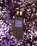 The image features a perfume bottle with a dark glass design set against a backdrop of purple leaves. The bottle has a rectangular shape with soft edges, and the cap has a gold finish with a rope-like texture.  The surrounding leaves create a rich, vibrant setting that emphasizes the bottle's elegance, suggesting that the perfume may have floral or botanical notes.