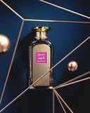 The image features a perfume bottle against a dark blue background, nestled within an artistic arrangement of golden geometric lines and spheres, creating a sophisticated and modern visual. The bottle has a textured glass design with a pattern of small, densely packed squares, and is capped with a gold-colored top embossed with a circular pattern. 