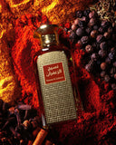 The image features a rectangular glass perfume bottle with a gold cap, set against a vivid background of deep reds and dark tones suggestive of spices. The bottle has a textured surface with a square pattern, and a maroon label with Arabic script reading "نسيج الزعفران" (Naseej Al Zafran), accompanied by the English transliteration. The backdrop includes richly colored spices like saffron and cloves, and cinnamon sticks, evoking the scent profile of the perfume and suggesting a warm, exotic fragrance.