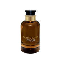 The image shows a cylindrical amber glass perfume bottle with a black cap and a gold band around the neck. The bottle has the text "NIGHT SHADOW" printed in large, capitalized, serif font in the center, with the brand name "zimaya" in a smaller, simple font below. The background is plain and white, highlighting the product.