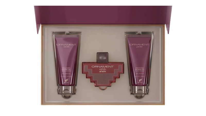 The image shows a gift set by Afnan Perfumes, "ORNAMENT purple allure". It features a perfume bottle and two tubes of shower gel, all in a coordinated deep purple color. The bottle has a unique, cross-shaped design with a red body and silver metalwork. The shower gels are labeled "ENERGIZING SHOWER GEL POUR FEMME" and are housed in sleek, metallic purple tubes.