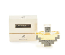 The image presents a perfume set which includes a bottle and packaging box. The perfume bottle has a unique, cross-like geometric design with clear and gold sections and a silver, mesh-patterned cap. The front of the bottle features the label "ORNAMENT" in a serif font with "pour femme" beneath it, indicating it's a women's fragrance from the brand AFNAN. The box behind the bottle is textured, primarily off-white with a gold band and the same branding as the bottle. 