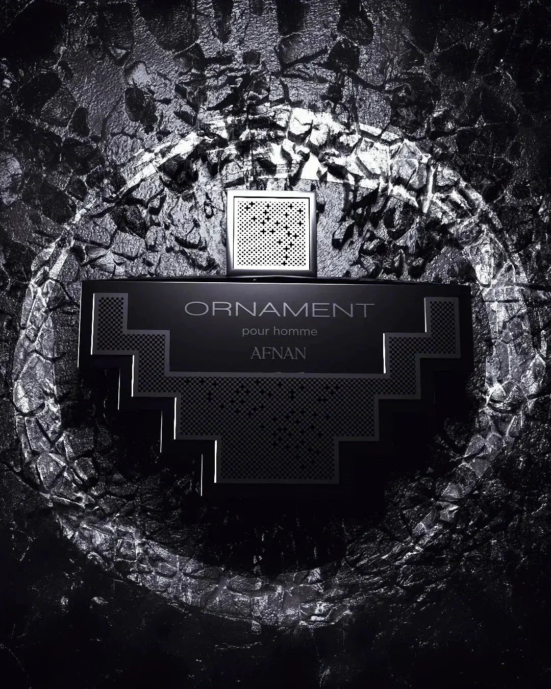 The image displays a monochrome perfume bottle with a distinctive geometric shape set against a textured, dark background resembling cracked stone or dried earth. The bottle is black with silver details and features a label with the text "ORNAMENT pour homme AFNAN," specifying that it is a men's fragrance from the brand AFNAN. The bottle cap has a mesh pattern, which adds to the overall modern and masculine aesthetic of the image. 