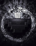 The image displays a monochrome perfume bottle with a distinctive geometric shape set against a textured, dark background resembling cracked stone or dried earth. The bottle is black with silver details and features a label with the text "ORNAMENT pour homme AFNAN," specifying that it is a men's fragrance from the brand AFNAN. The bottle cap has a mesh pattern, which adds to the overall modern and masculine aesthetic of the image. 