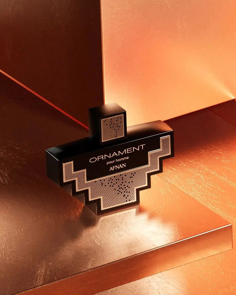 The image features a black geometric perfume bottle with silver accents and a mesh pattern on the cap, situated on a reflective surface with a warm, copper-toned backdrop. The bottle displays the label "ORNAMENT pour homme AFNAN," indicating that it is a men's fragrance from the brand AFNAN. The lighting casts soft highlights and shadows across the scene, enhancing the bottle's angular design and creating an atmosphere of sophisticated luxury.