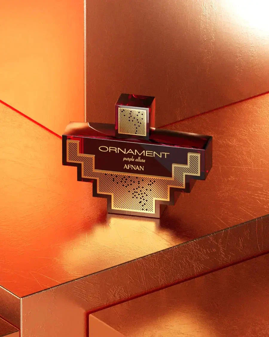 The image features a creatively designed perfume bottle from Afnan Perfumes. The bottle has a cross-like silhouette with a tiered, geometric shape and is predominantly dark red to black in color. It has a detailed gold pattern over a grid texture in the central section, giving it a luxurious appearance.  The lighting creates a warm, glowing effect on the metallic surfaces, enhancing the opulence of the product presentation.