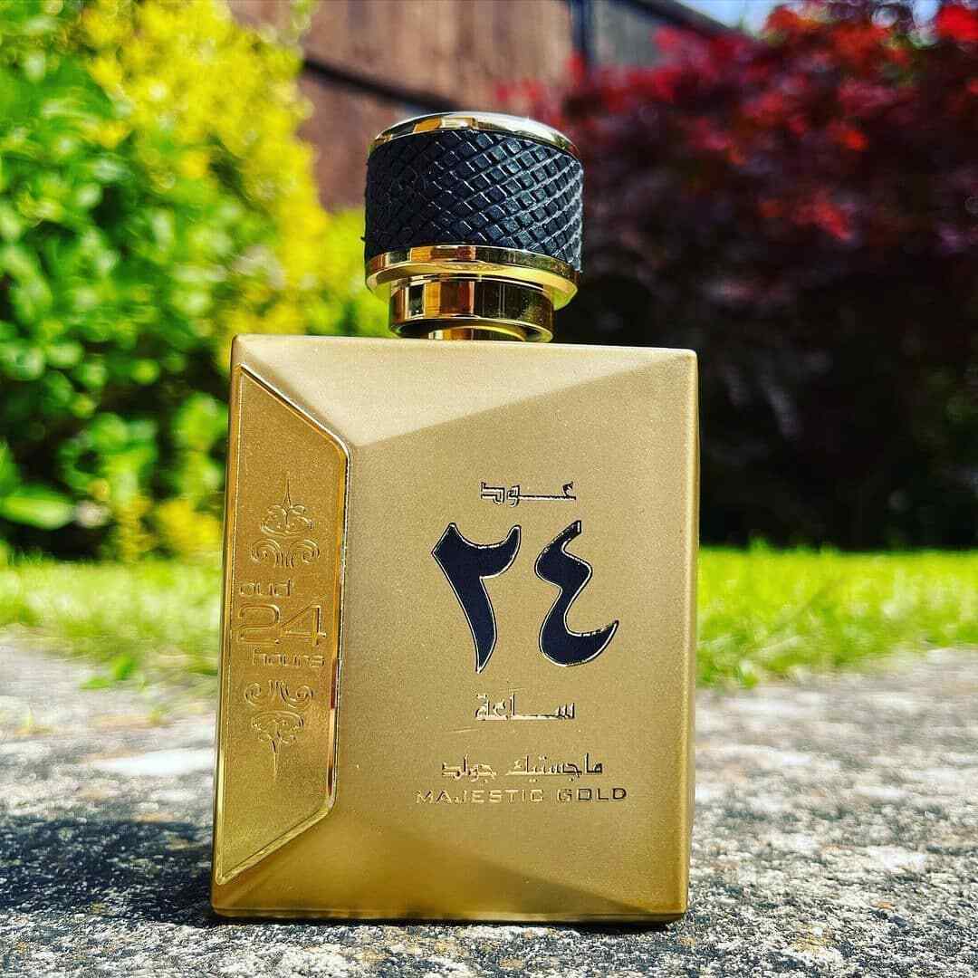 The image displays a golden perfume bottle with a textured black cap, placed on a concrete surface with a background of lush greenery and a red-leaved tree. The bottle features Arabic calligraphy in black and the English phrase "MAJESTIC GOLD" towards the bottom. The bottle is positioned in natural lighting, highlighting its metallic sheen and the elegant embossed details on its side. The outdoor setting gives the image a fresh and vibrant ambiance.