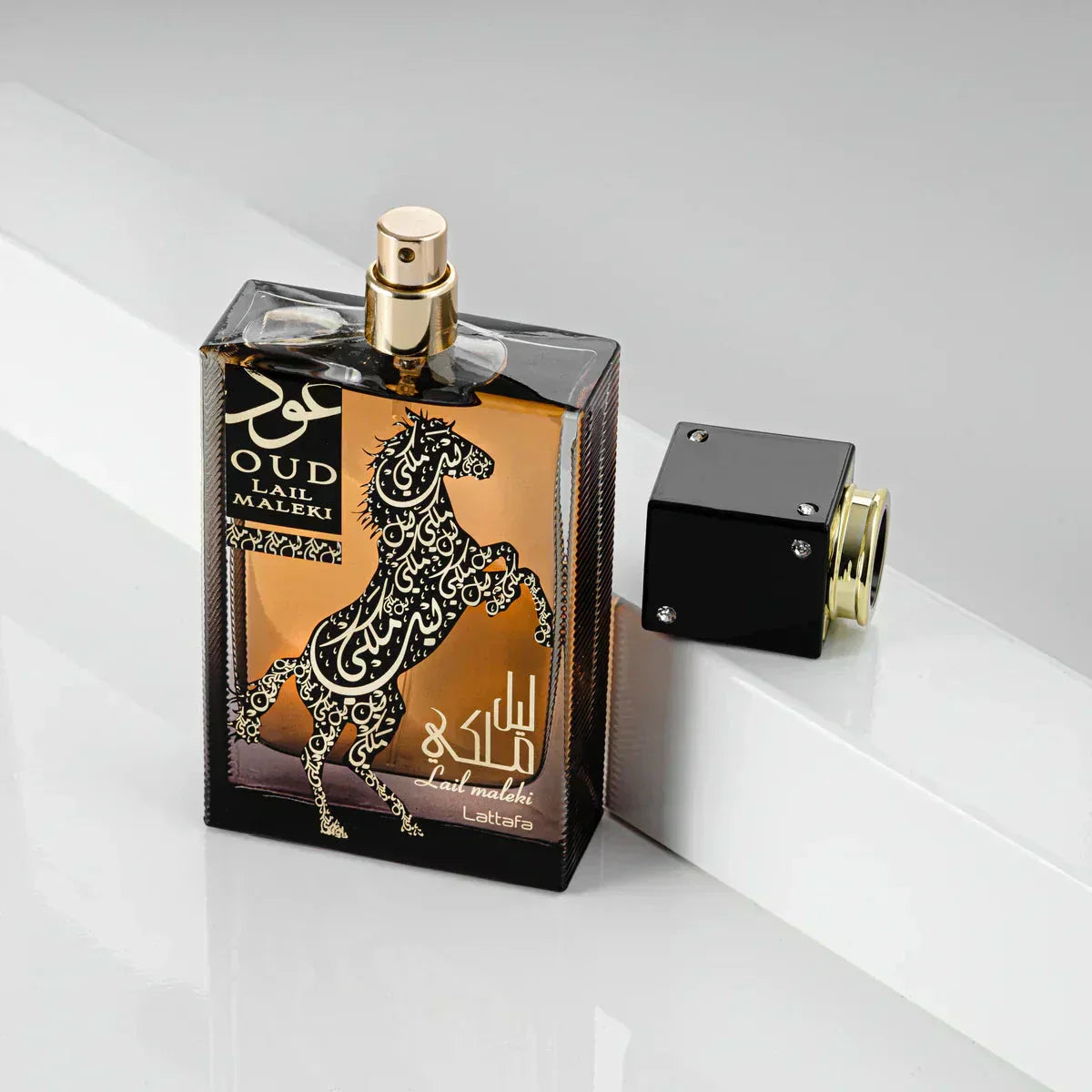 The image presents "Oud Lail Maleki" by Lattafa, a perfume bottle with a unique design. The bottle has a dark, amber glass with a rearing horse and Arabic calligraphy in white and gold, creating an ornate contrast. The name of the fragrance is written in both Arabic and English. The cap of the bottle is black with gold trim and appears to be detached, revealing a gold-colored spray nozzle. This perfume is presented on a reflective surface with a white backdrop, adding to the luxurious feel of the product.