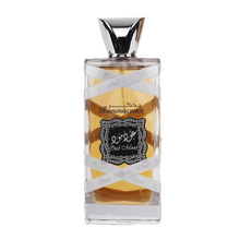 The image displays a perfume bottle with the following attributes:  The bottle is rectangular with a clear glass design, allowing a view of the amber-colored perfume inside. A silver ribbon with the words "Oud Mood" printed in a repeating pattern wraps around the bottle. The central label is ornate with a black background and white decorative border; it features Arabic calligraphy and the word "Reminiscence" in English script.
