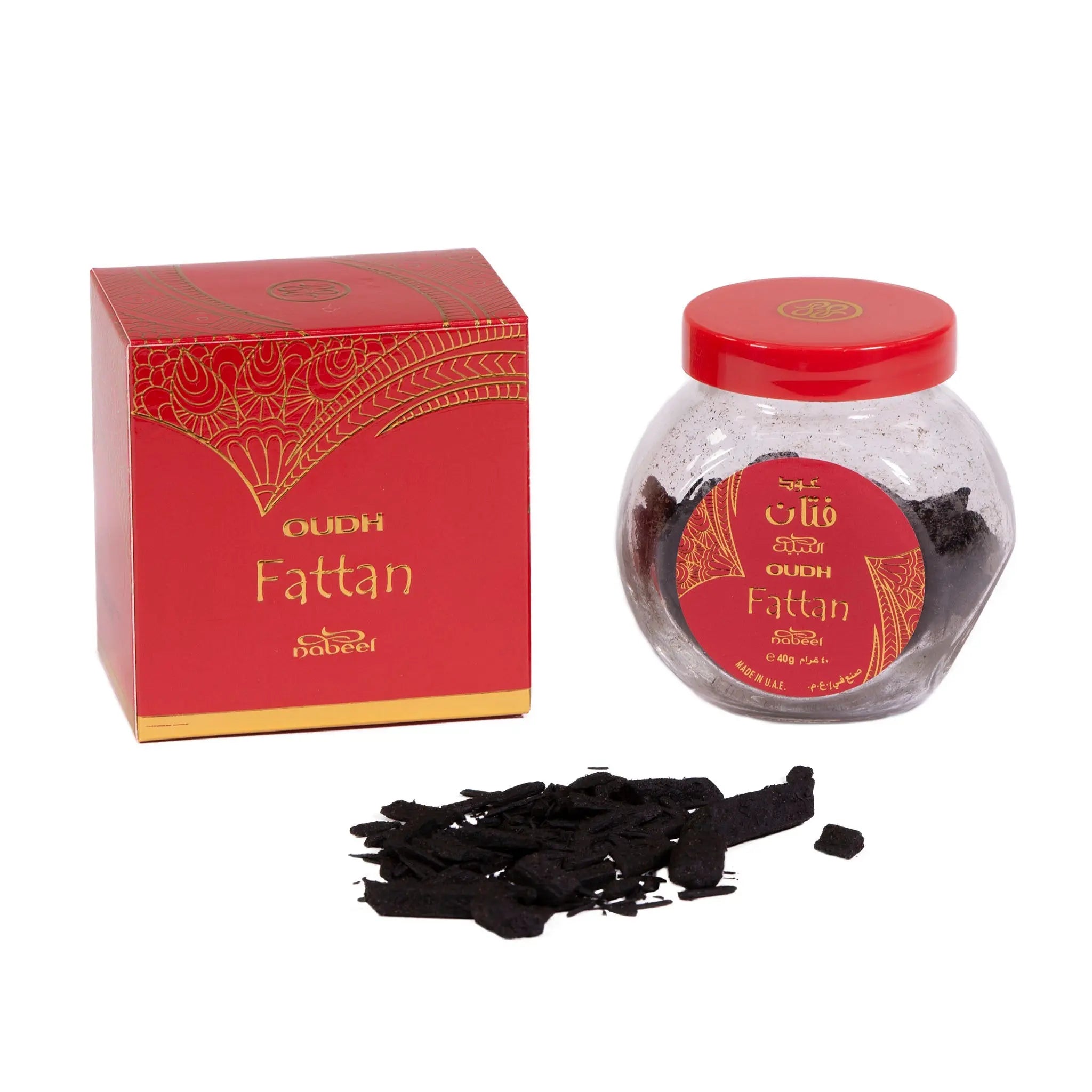 The image shows a product called "OUDH Fattan" by Nabeel, presented with a red and gold box and a clear glass jar with a red lid. Both the box and jar feature an intricate, traditional Arabic-style golden pattern. Arabic and English text on the label identify the product. In front of the jar, there are several pieces of dark, resinous material, presumed to be oudh (agarwood), used for its aromatic properties.