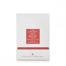 The image displays a white packaging box with a red label for a fragrance product. The label includes the name "PEACH MUSK" in English, with corresponding Arabic script above it. Below, it states "IBRAHEEM AL.QURASHI SPECIAL KHAMRIYA FOR HAIR." A red strip at the box's base matches the label, and the brand's logo is printed in red at the bottom.