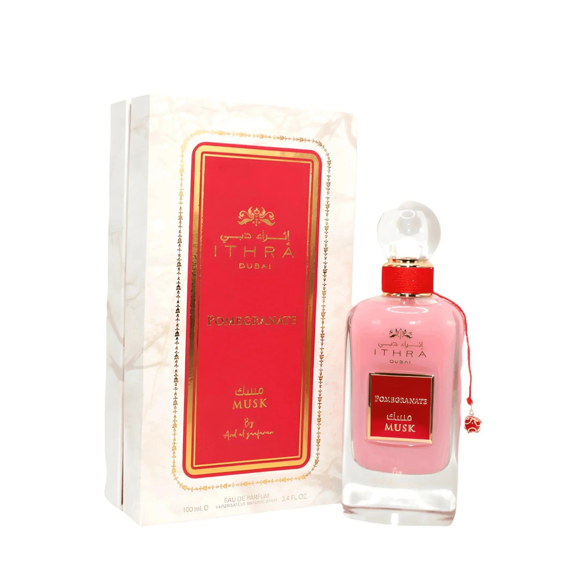 This image shows a bottle of Ithra Dubai Pomegranate Musk perfume next to its packaging. The bottle has a clear glass design with a pink hue and a white gradient base, topped with a round glass cap and a gold-threaded neck. A small gold charm designed like a pomegranate is attached to the neck with a red string.  The box has a marble-like design with gold and red accents, and it features the brand name Ithra Dubai and the fragrance name Pomegranate Musk in both Arabic and English script.