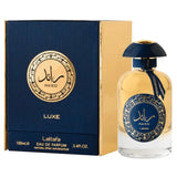 The image features a perfume bottle and its packaging:  The perfume bottle has a clear glass body with a golden-yellow liquid, and a navy blue dome-shaped cap. A navy blue ribbon is tied around the neck of the bottle, complementing the cap. The front of the bottle displays a navy blue label with gold decorative patterns and Arabic calligraphy of the name "RA'ED," with the brand "Lattafa" beneath it. The packaging next to the bottle is a box with a navy blue and gold color scheme. 