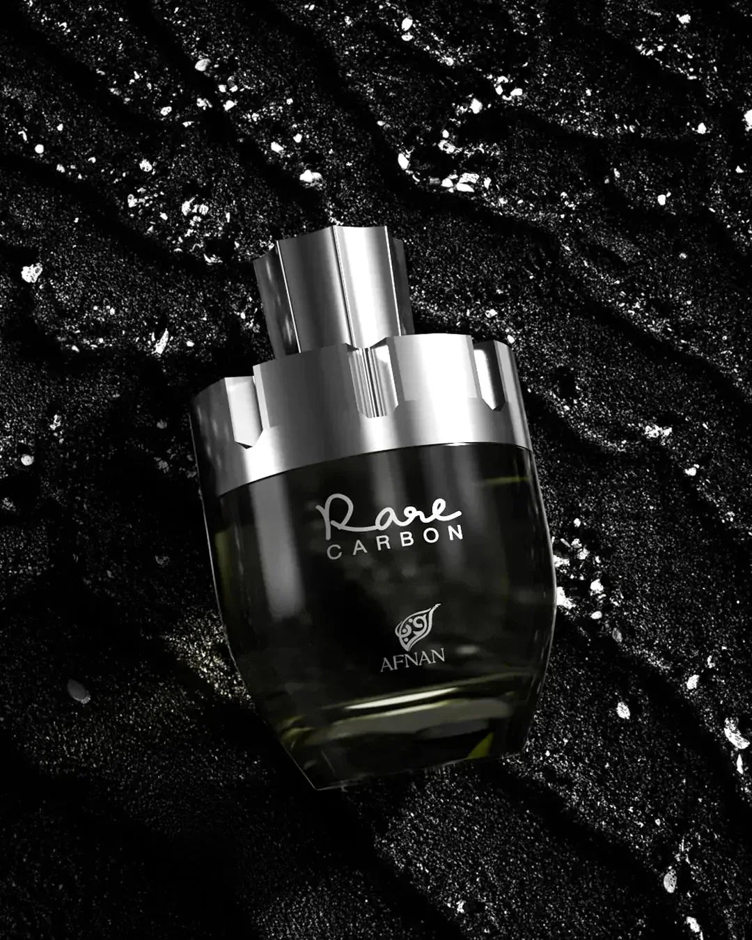 The image depicts a perfume bottle from the brand AFNAN, specifically the "Rare CARBON" fragrance. The bottle has a sleek, modern design with a transparent glass body that fades from clear to a dark gradient at the base, suggesting the color of the perfume within. The bottle is placed against a textured black background with sparkling silver flecks scattered throughout, giving the impression of a luxurious and elegant product. 