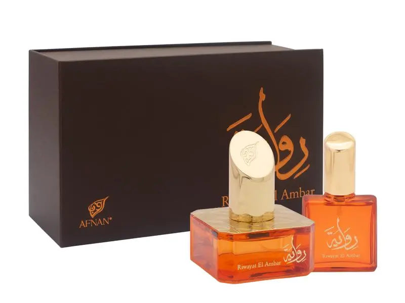 The image shows a set of two perfume bottles with a dark brown backdrop provided by their box packaging. On the left, there is a larger rectangular packaging box with the brand logo "AFNAN" in gold at the lower left corner and an elaborate calligraphic design in gold at the center. The design possibly represents the name of the perfume in Arabic script. In front of the box, there are two transparent orange-colored glass perfume bottles, each with gold caps.